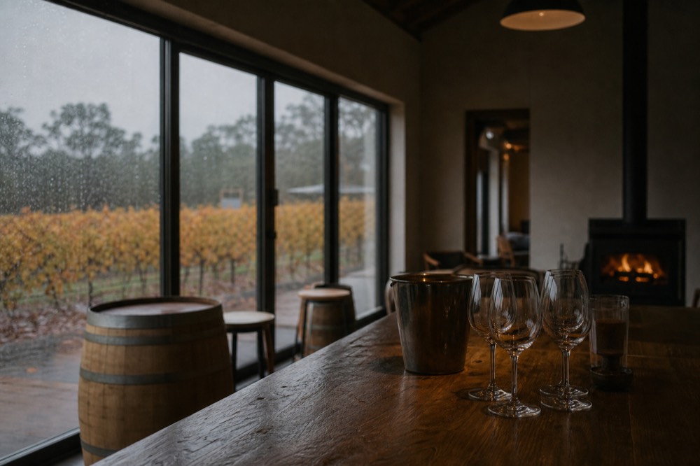 A near-empty tasting room in soft winter light, timber counter and a fireplace glowing in the background, rain on the window, no branding or people.