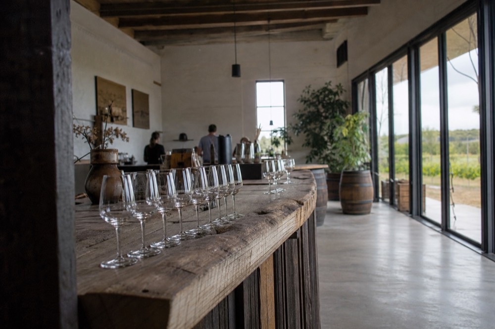 An interior of a rustic cellar door with a long timber tasting bar, rows of glasses set out, warm daylight through tall windows, no branding or people.