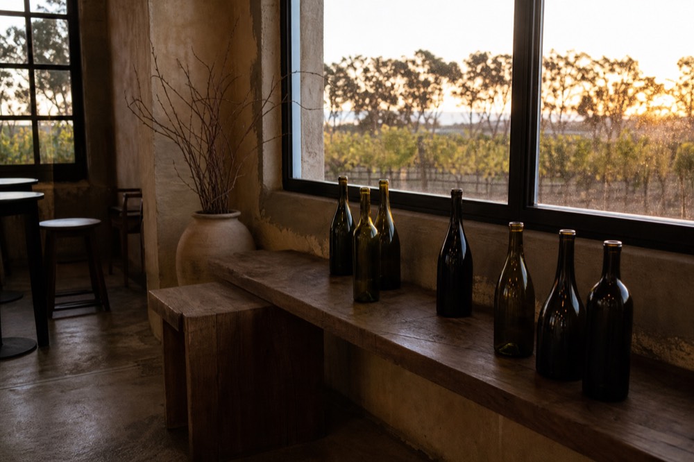 A small boutique tasting room interior with a handful of unlabelled bottles on a rustic bench, warm light through a window, no branding or people.