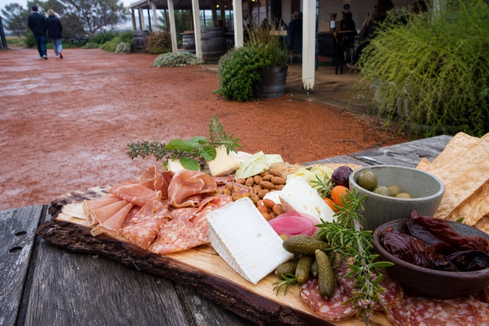 A grazing platter of local produce, cured items, cheeses and native-looking herbs on a rustic timber board, outdoor daylight, no text or branding.
