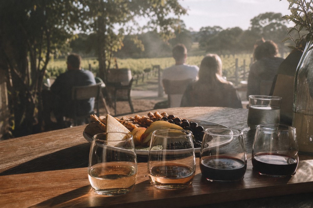 A close view of small tasting pours and a plate of local produce on a timber table outdoors, soft natural light, no logos or text visible.