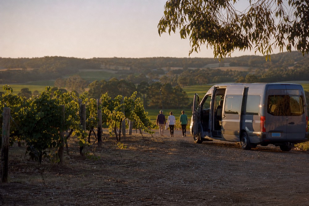 A small tour van on a gravel drive beside vineyard rows, doors open, green valley beyond, clear afternoon light, no branding or number plates.