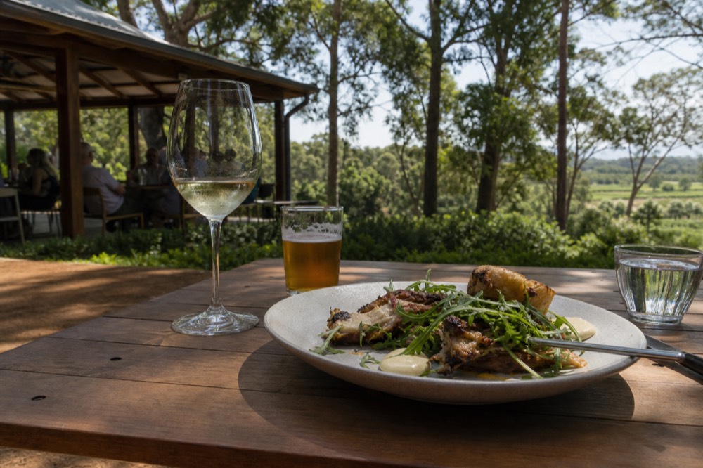 A gourmet lunch plate beside a glass of wine and a small beer on a timber table at a winery, greenery beyond, soft midday light, no branding.