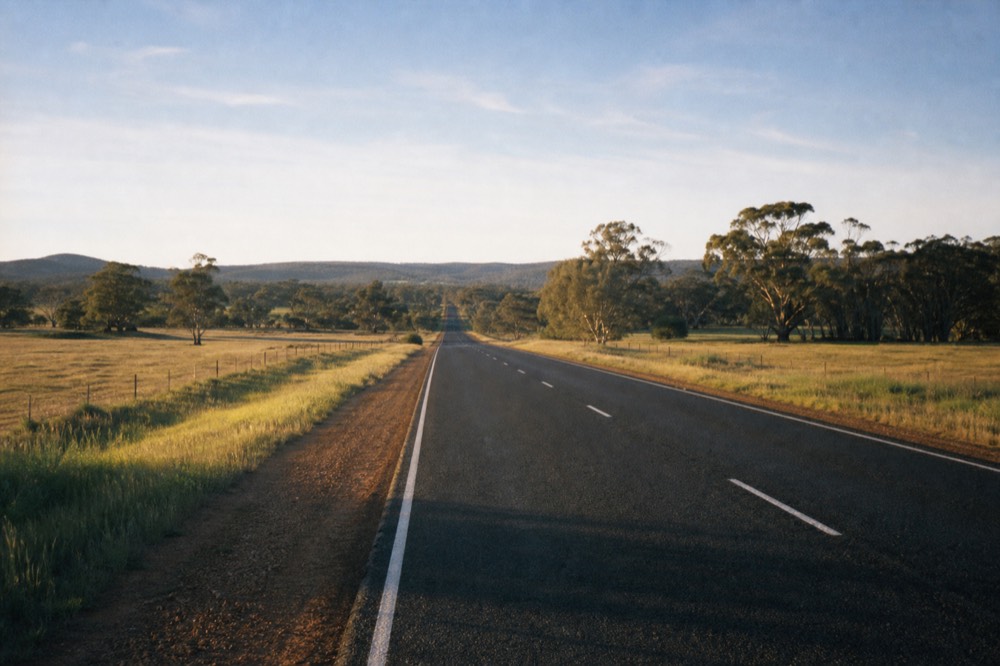 A long straight regional highway running through green pasture and stands of eucalypt under a broad morning sky, gentle hills on the horizon, no traffic or signage.